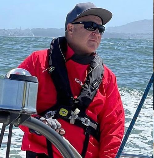 Brendan is driving a boat off the shore of Santa Monica Bay. He is wearing a red jacket and a PFD. He has a serious look on his face as he drives the boat.