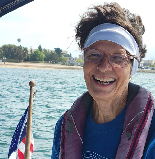 Debbie has a huge smile. The ocean and a beach are behind her. The wind is blowing her hair.
