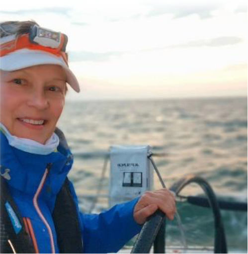 Leslie is smiling as she is steering a boat. The ocean is behind her. She is wearing a visor and a night lamp on her head.