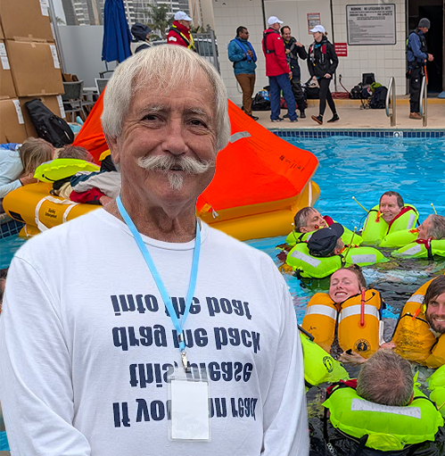Dave Roberts standing in front of DRYC pool with sailors in the pool all wearing lifejackets. A life raft is behind Dave. Dave has a nice handlebar mustache.