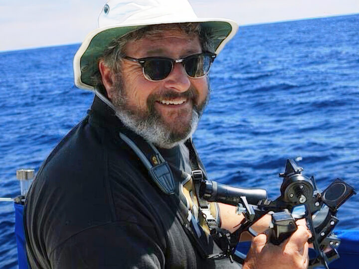 Capt. Mike Blecher on the ocean holding his sextant. He has on a floppy hat and sunglasses.