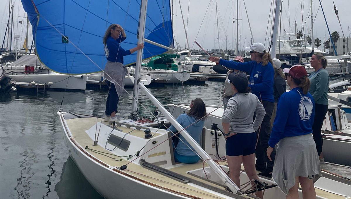 One woman learns bow and another learns pit position at the dock, with the spinnaker flying, and the rest looking on