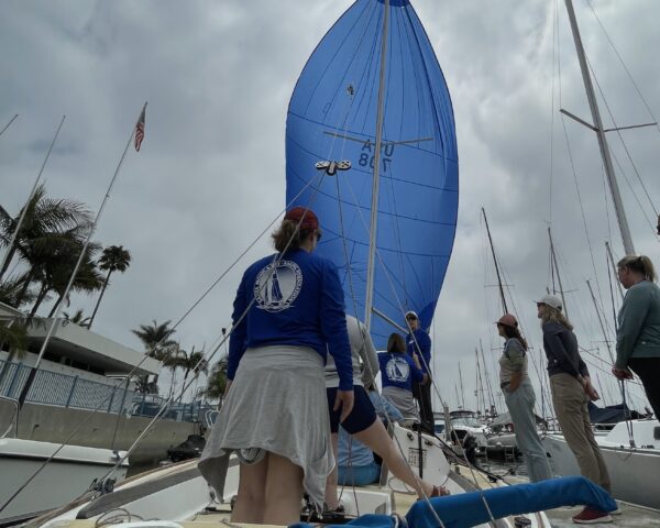 A group of people in and around a boat with the spinnaker flying, learning about spinnaker trim from the instructor.