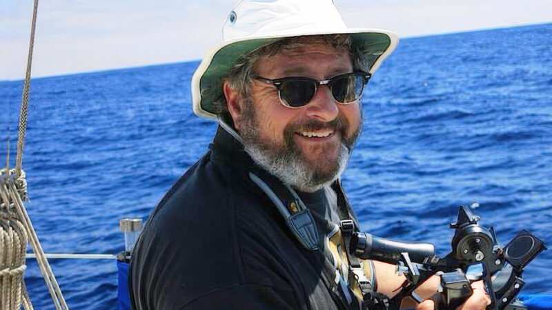 Capt. Mike Blecher on the ocean holding his sextant. He has on a floppy hat and sunglasses.