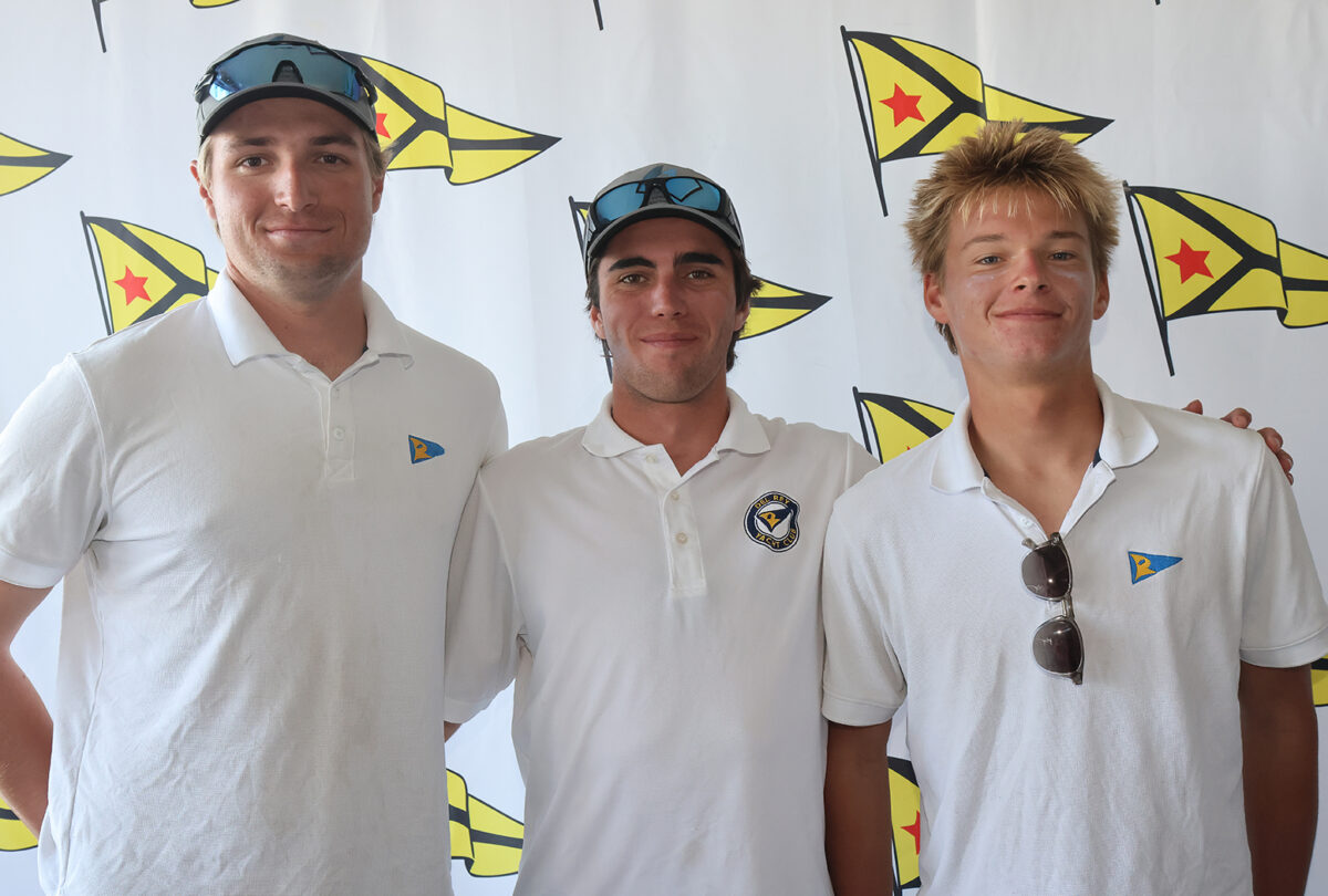 Three proud sailing race competitors (young men) in matching white shirts standing in front of a step-and-repeat with the Del Rey Yacht Club logo on it