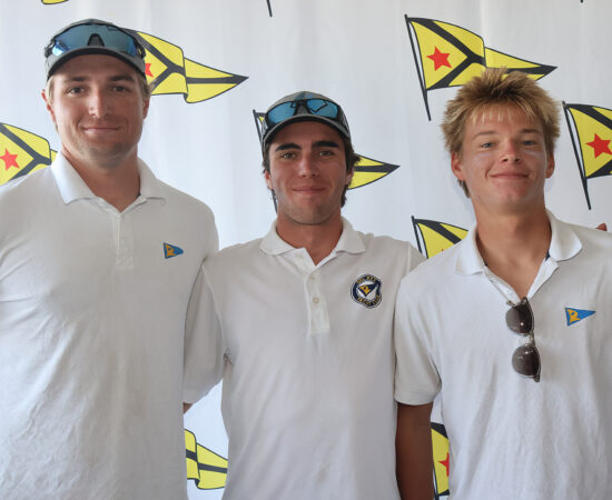 Three proud sailing race competitors (young men) in matching white shirts standing in front of a step-and-repeat with the Del Rey Yacht Club logo on it